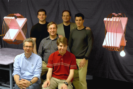 The wireless power transmission research team standing in a lab between two styrofoam frames wrapped in copper wire. From the right frame and coil hangs a shining lightbulb.